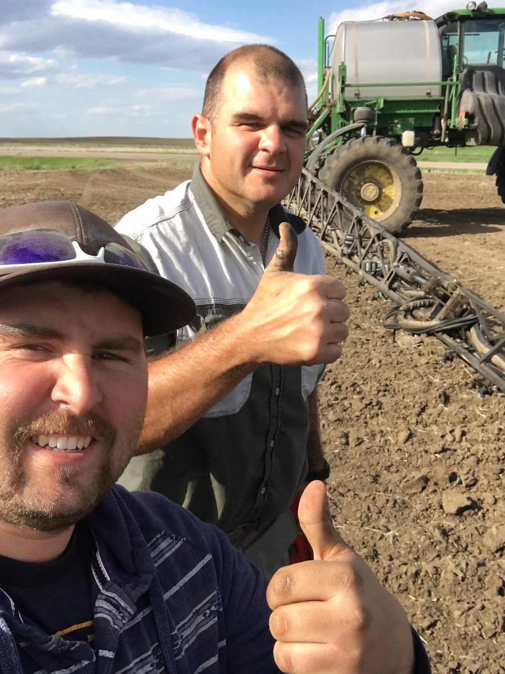 Two people giving the thumbs up in a field with a tractor behind it